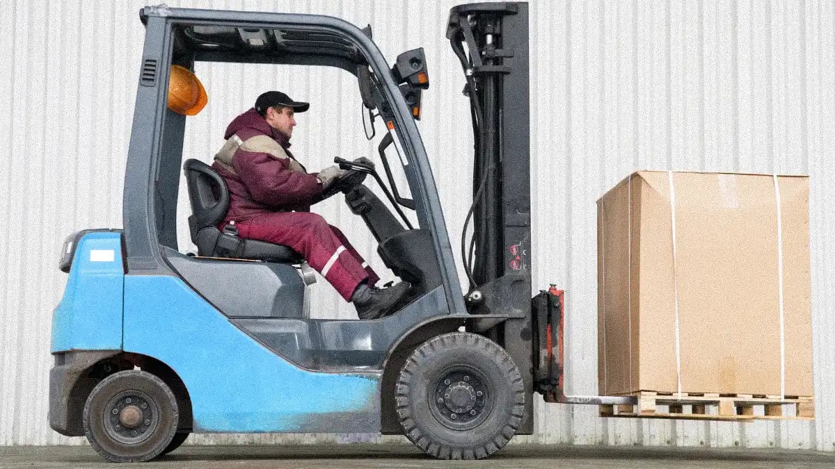 A restoration contractor operating a blue forklift, lifting a large cardboard box on a pallet in an outdoor industrial setting.