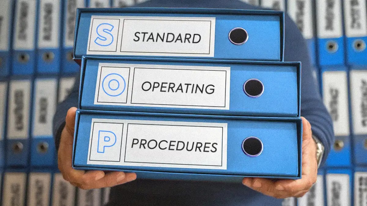 A restorer holding three blue binders labeled ‘Standard Operating Procedures’ (SOP) in front of a shelf filled with similar binders on water damage, fire, initial assessment and other workflows.