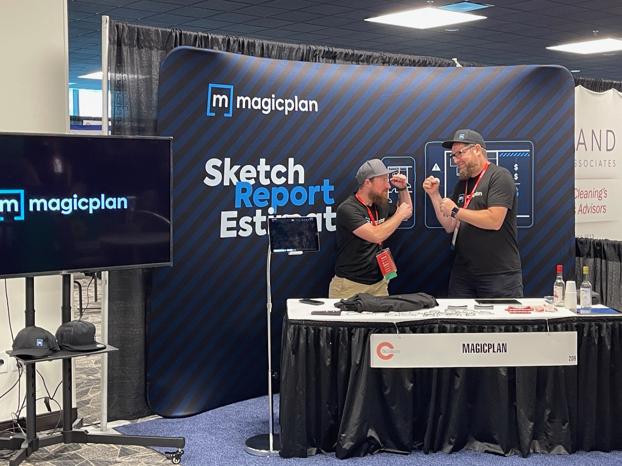 Josh Winton from Discreet Restoration and Allan Bassenden in a Restoration Industry Trade Show Behind magicplan's Booth Table Fighting. TV with the magicplan logo and banner.
